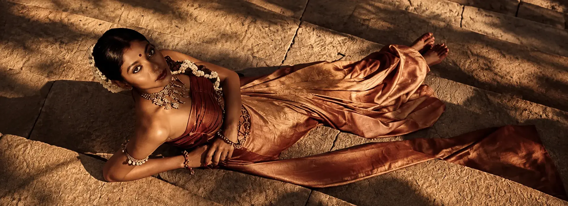 A woman reclining on warm stone in a copper silk drape, wearing traditional jewellery, lit by low evening sun